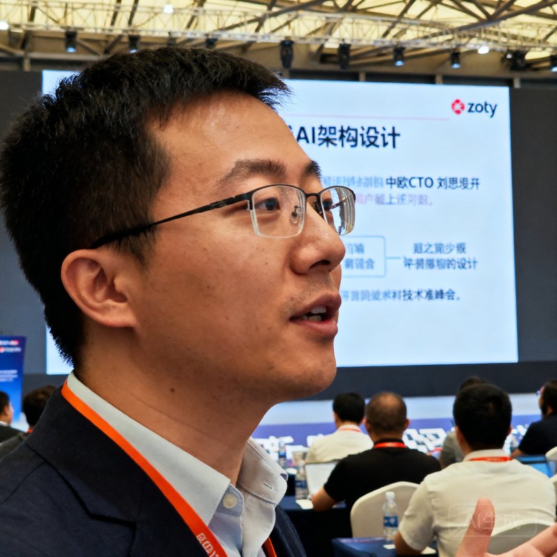 zoty中欧CTO刘思远在技术峰会上演讲的近景照片，他佩戴眼镜，正在大屏幕前讲解AI架构设计